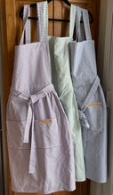 Load image into Gallery viewer, Purple, green and blue striped Hovden Wear aprons hanging on a kitchen cabinet.
