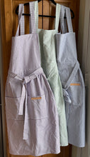 Load image into Gallery viewer, Purple, green and blue striped Hovden Wear aprons hanging on a kitchen cabinet.